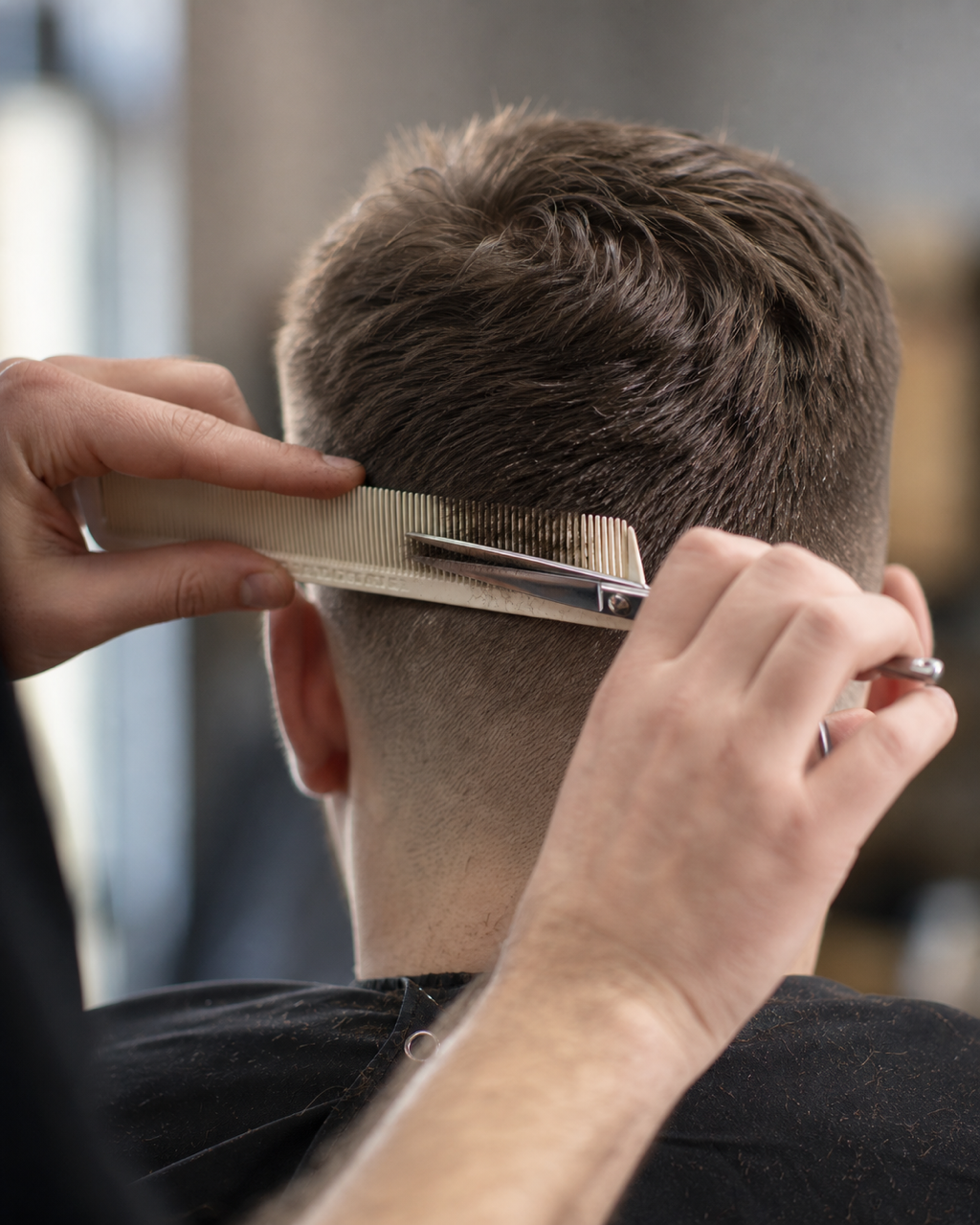 A barber's hands guiding scissors and comb through the back of a short haircut